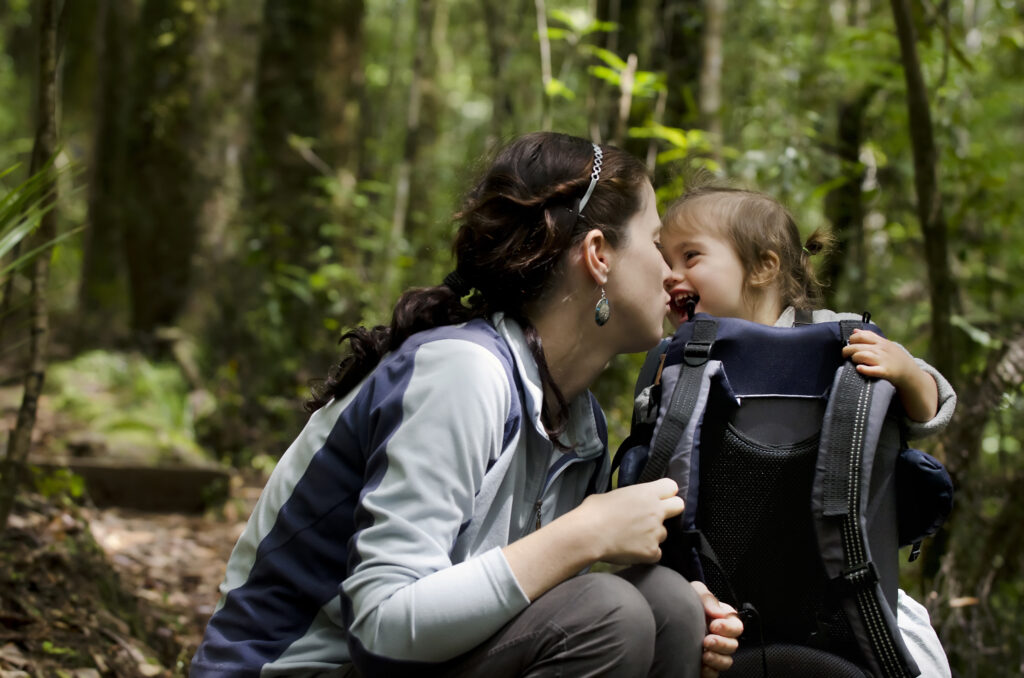 A mother and her baby backpacking through a forest, show their love for one another while they stop for a rest.